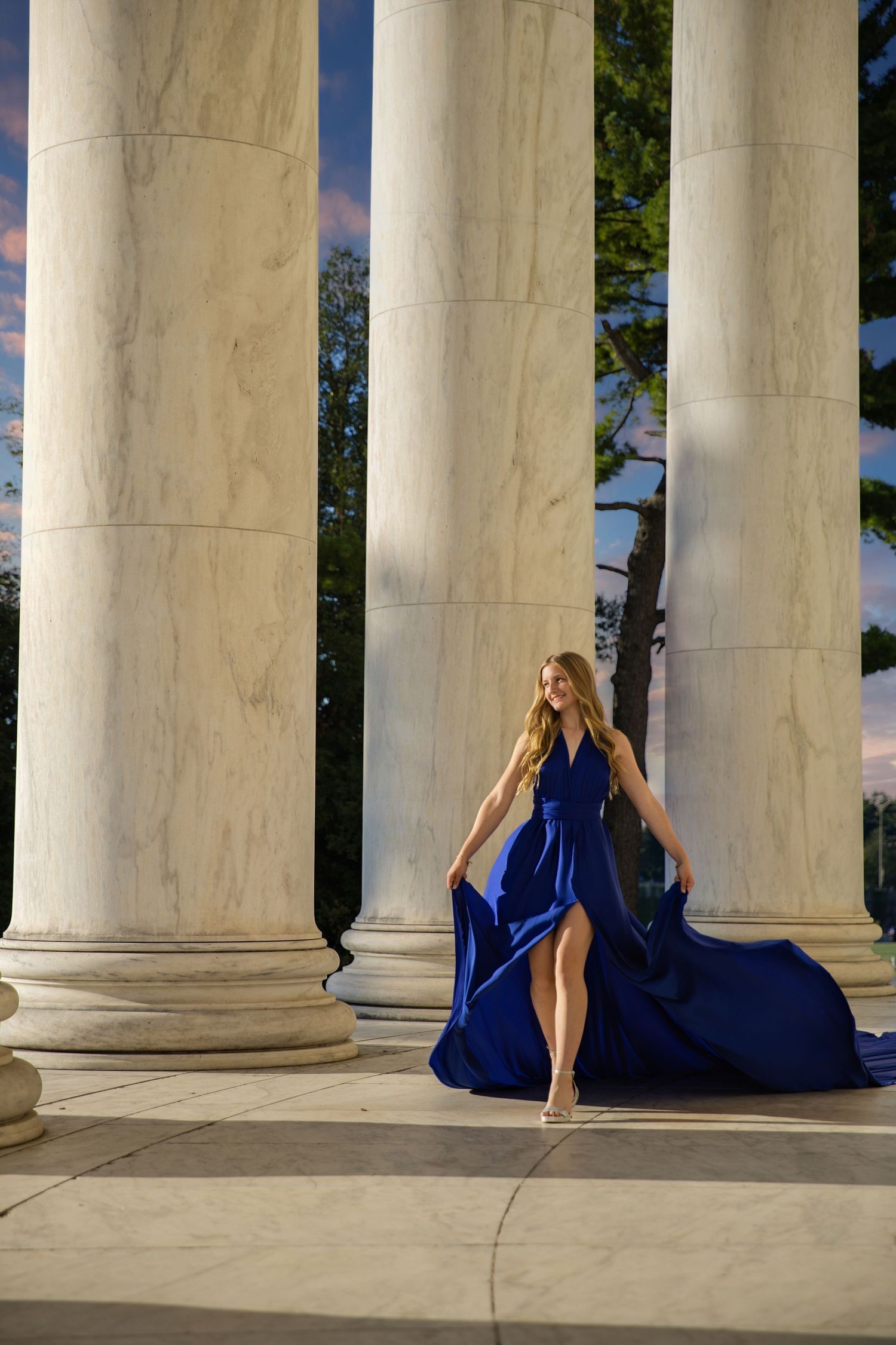 Woman in flowing blue dress poses elegantly between classical white stone columns in artistic photo series. Flying Dress Photoshoot