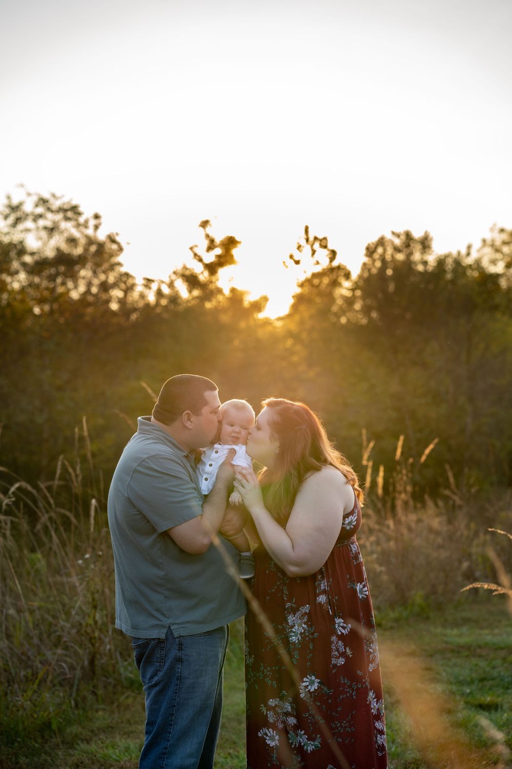 Family portraits with mom and dad holding baby son in between them each kissing one side of his cheeks, standing in a field with trees.
