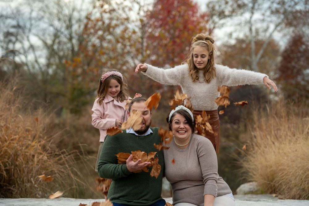 Fall family portrait of Mom and Dad sitting on a flat rock bridge while their two daughters throw leaves on them from behind.