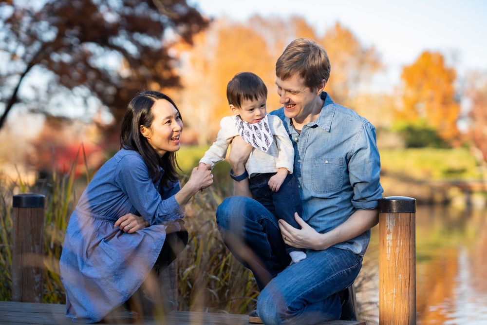 A fall family portrait of mom, dad and toddler son on a walkway over a lake at a garden.