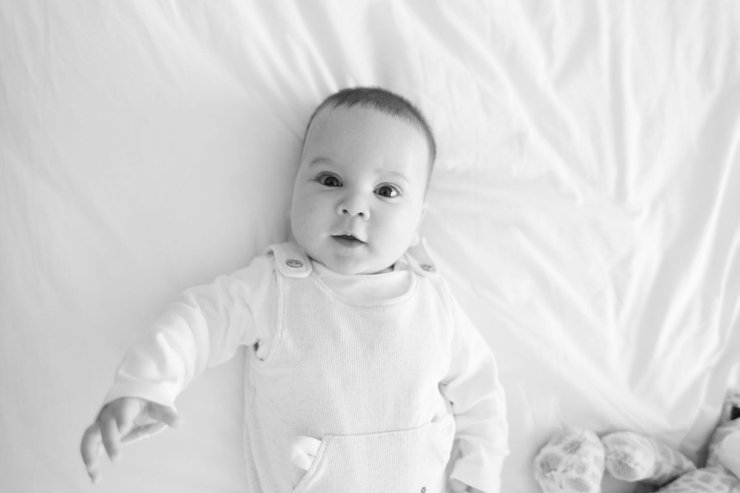 A sweet baby wearing white lies on cozy white bedding while looking up at the camera with bright eyes.