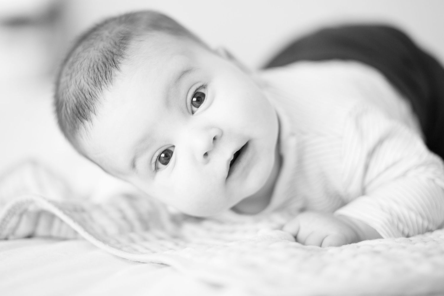 Black and white portrait of a baby lying on white bedding looking up with bright eyes and a gentle expression.