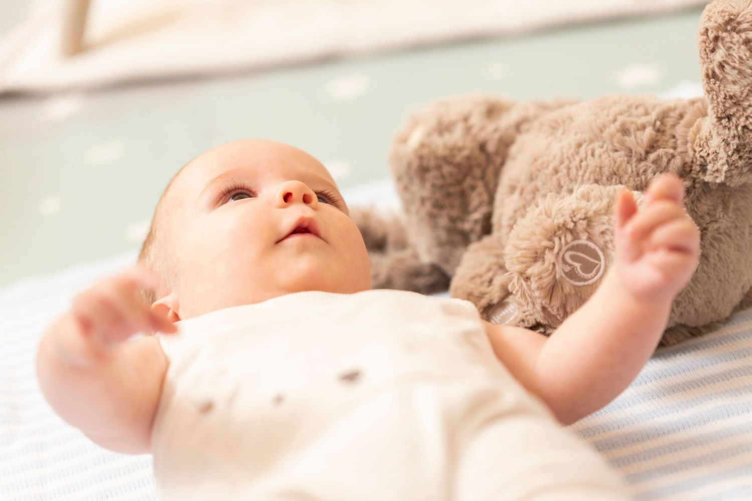 Infant lying on soft white bedding with a colorful toy visible in the background.