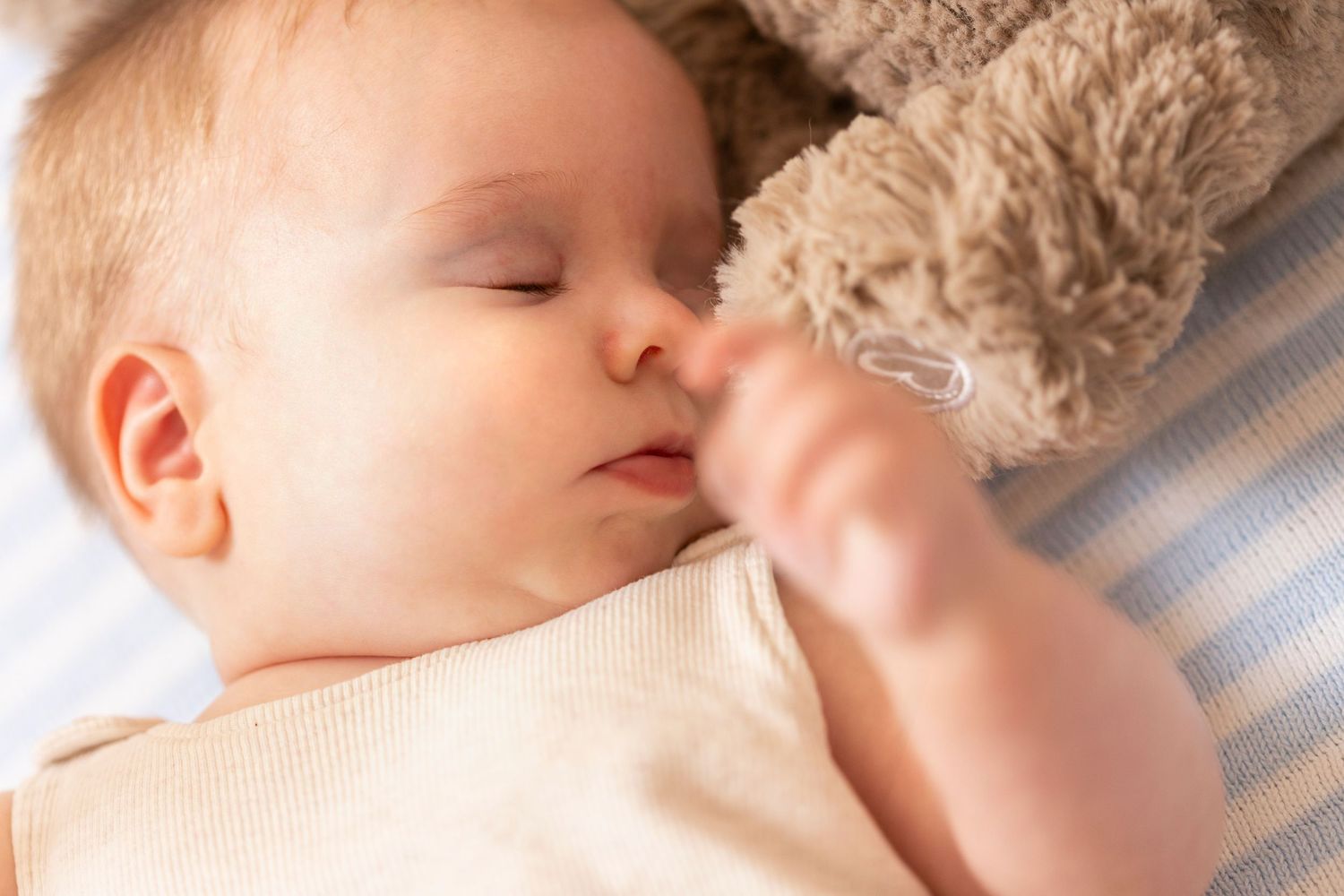 Sleeping baby cuddles with soft plush toy on striped bedding.