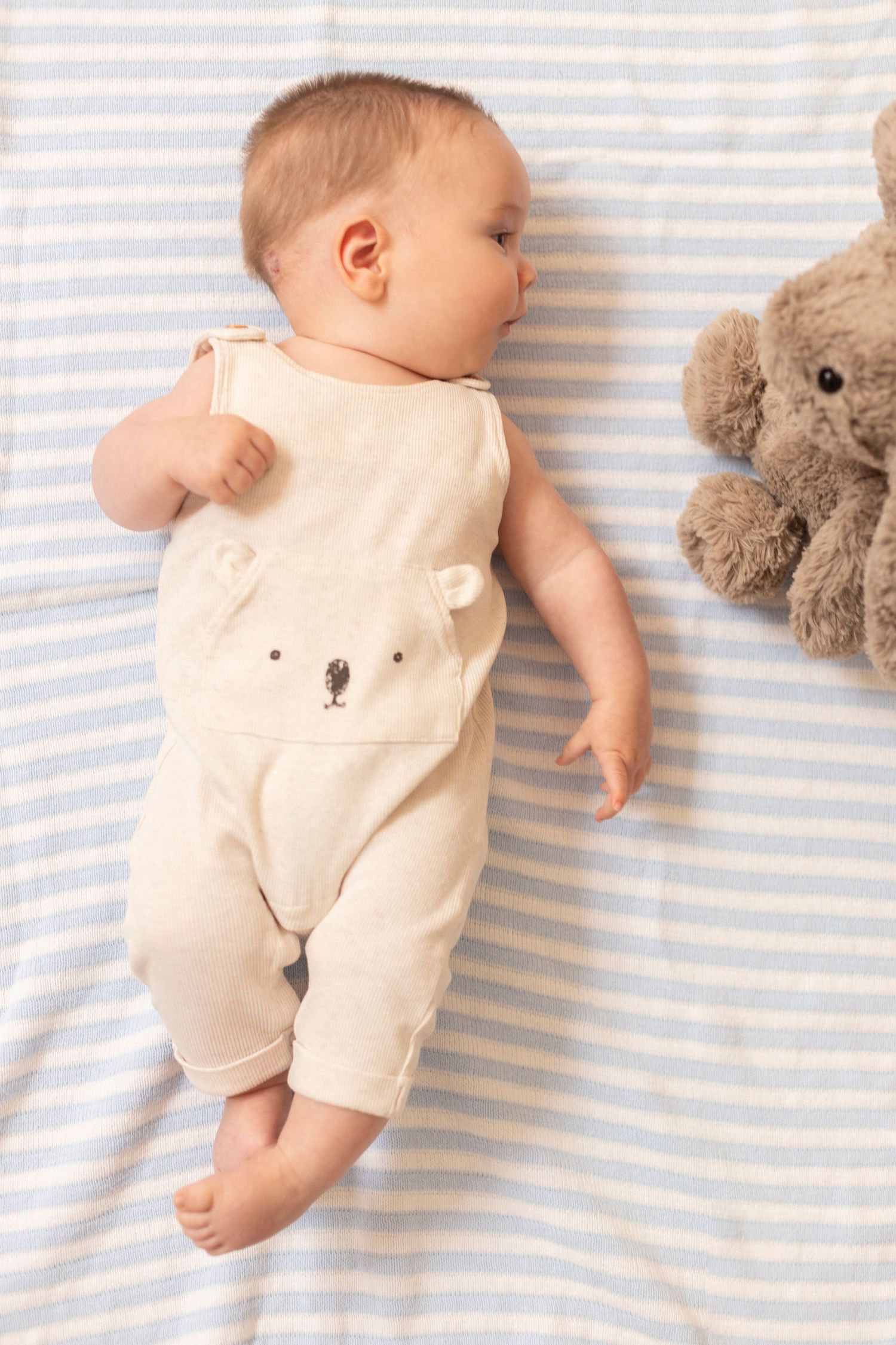 Baby in beige bear onesie rests on blue and white striped blanket beside stuffed bunny.