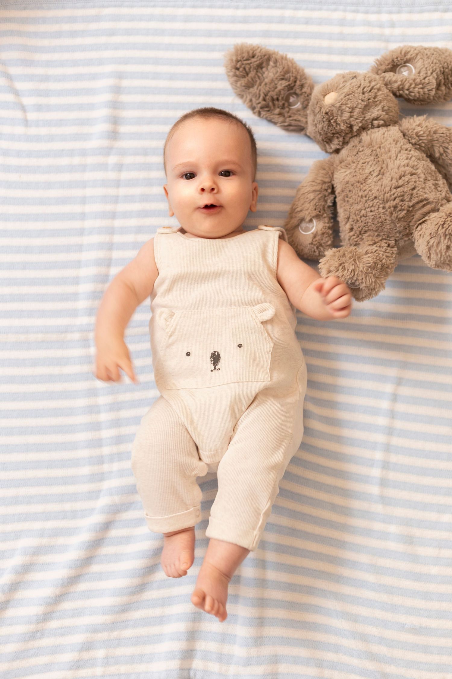 Baby wearing cream-colored bear romper lies on striped bedding next to plush bunny toy.