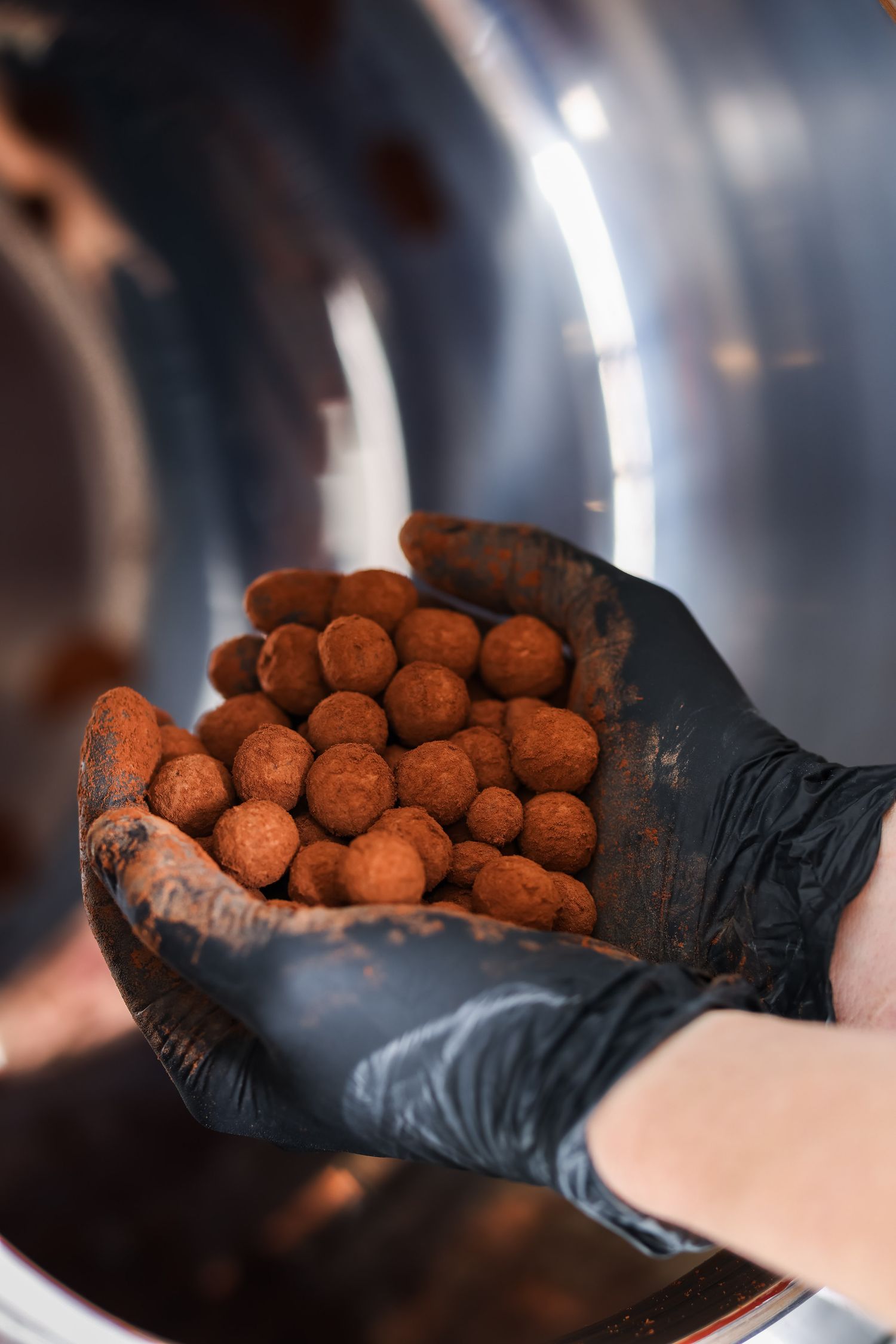 A hand wearing a black glove holds chocolate truffles dusted with cocoa powder against a dark background.