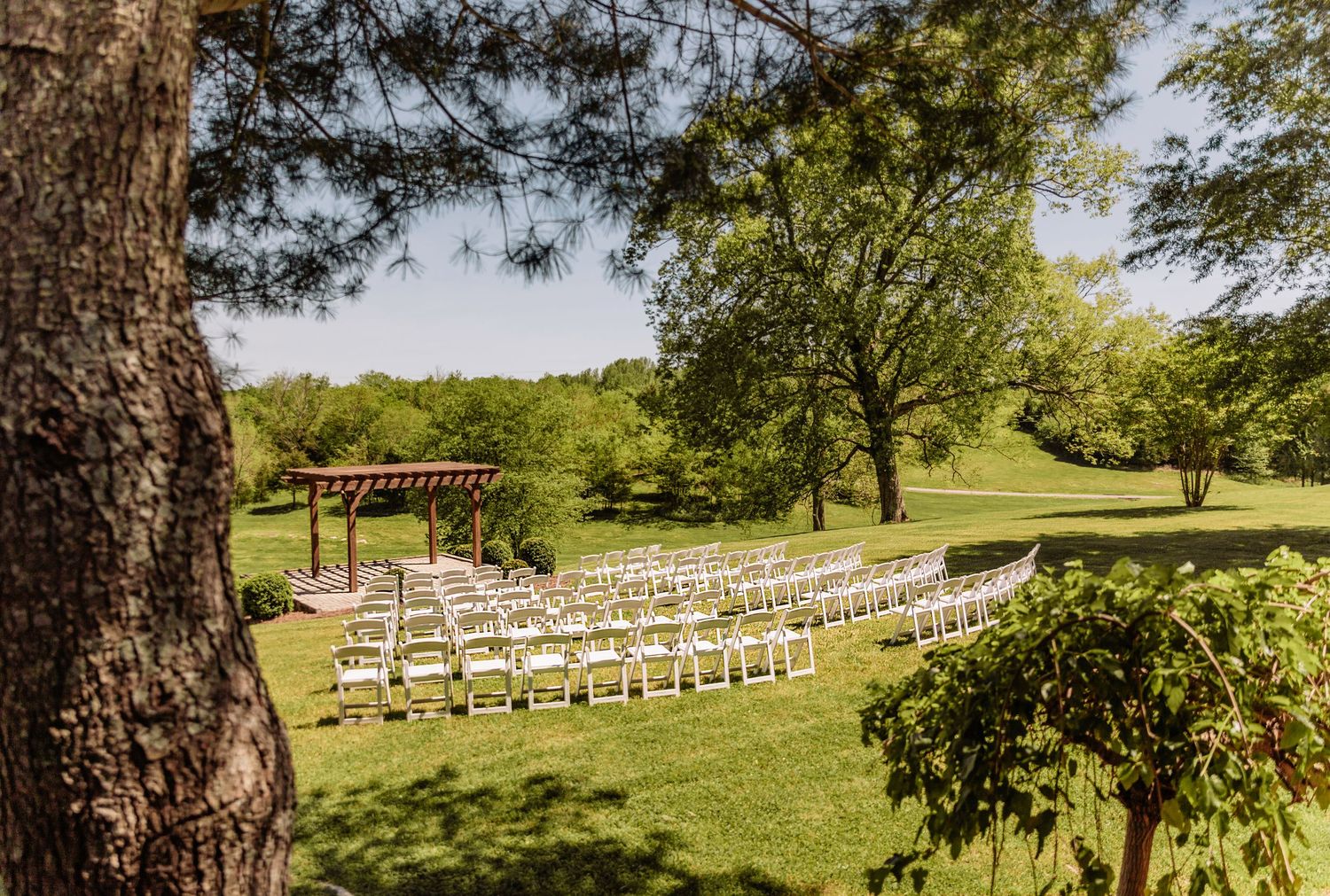 Outdoor wedding ceremony setup with white chairs arranged on a green lawn surrounded by tall trees.