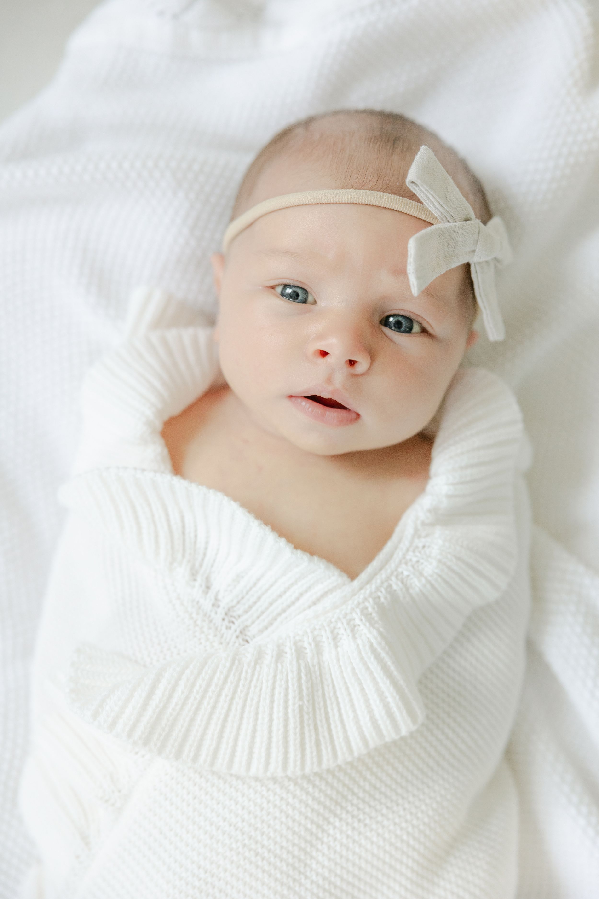 Sweet baby wearing white knit sweater lying on soft white bedding with delicate bow hair accessory. Hartford Newborn Photography Studio.
