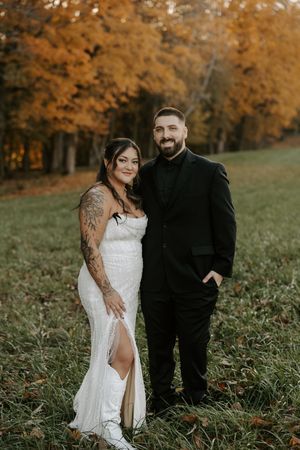 Autumn wedding portrait with couple standing in field with orange fall foliage behind them.