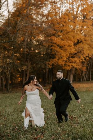 Wedding couple runs playfully through autumn field with orange and gold trees in background.