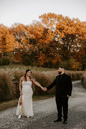 Wedding couple walks hand in hand on gravel path with golden autumn trees in background.