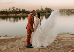 Wedding couple embracing by lakeside with flowing white veil in evening light.
