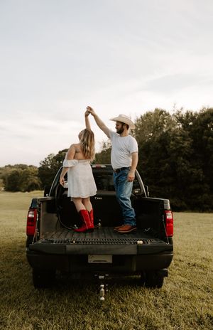 A couple sits casually in the bed of a black pickup truck parked in a grassy field.