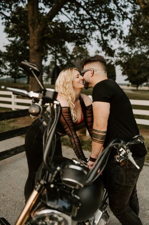 Couple poses intimately with a black motorcycle on a tree-lined path.