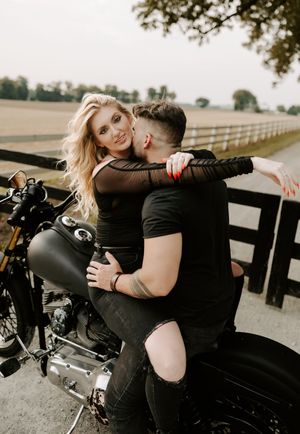 Romantic couple in black clothing poses with a motorcycle against rustic fence backdrop.