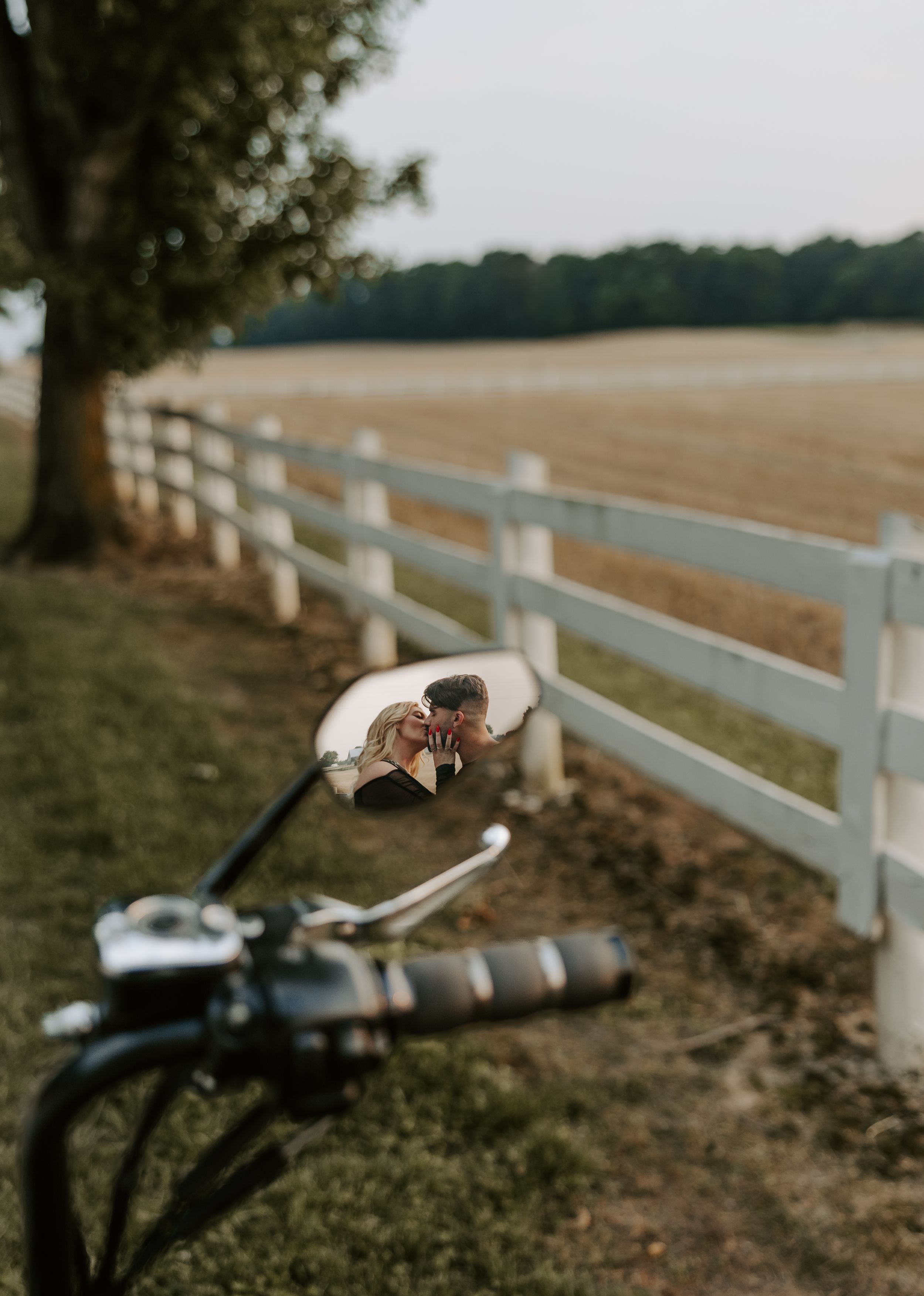 A motorcycle mirror reflection against a white fence and open field backdrop.