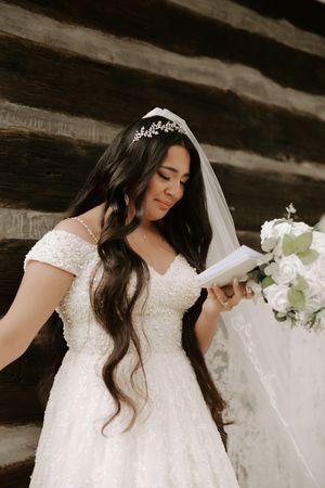 Beautiful bride in a white lace gown with flowing veil poses against a rustic wooden backdrop.