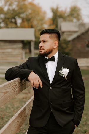 A groom in a black tuxedo and white boutonniere poses against a wooden fence during autumn.