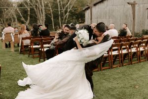 A flowing white wedding dress train trails behind on a grassy lawn with wooden chairs set up for an outdoor ceremony.
