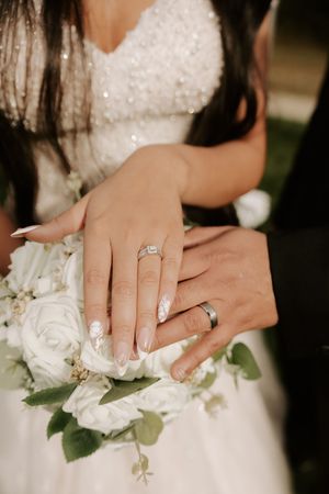 Close-up of newlywed couple's hands with wedding rings resting on white rose bouquet showing sparkling engagement ring.