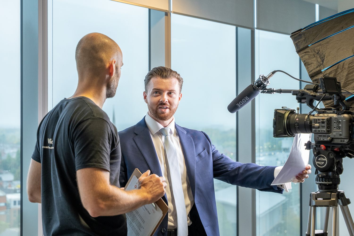 A professional video production crew films an interview in a modern office setting with city views through windows.