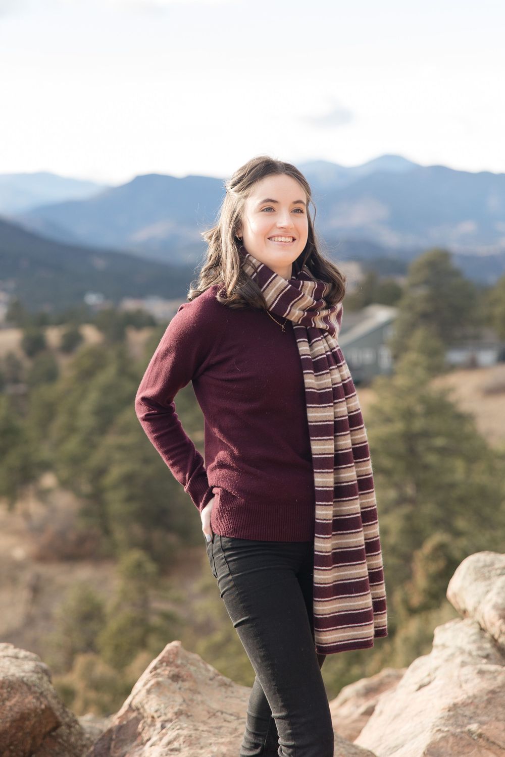 Person in burgundy sweater and striped scarf poses on rocky overlook with mountain vista in background.