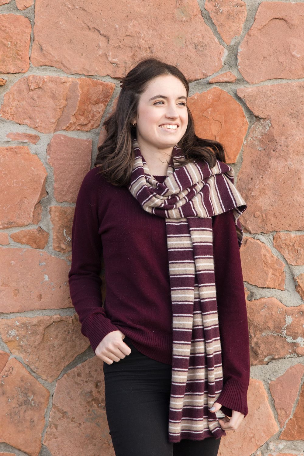 A person in a burgundy sweater and striped scarf poses against a rustic stone wall.