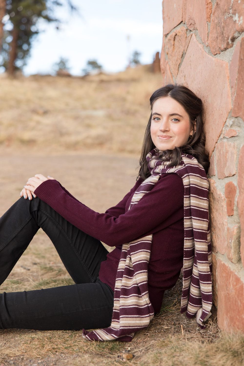 Casual winter outfit with burgundy sweater and striped scarf while sitting by stone wall outdoors.