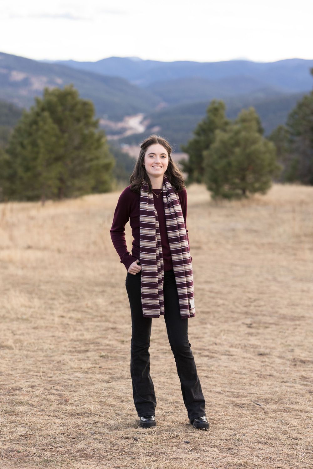 Person poses outdoors in a burgundy sweater and striped scarf against a mountainous landscape.
