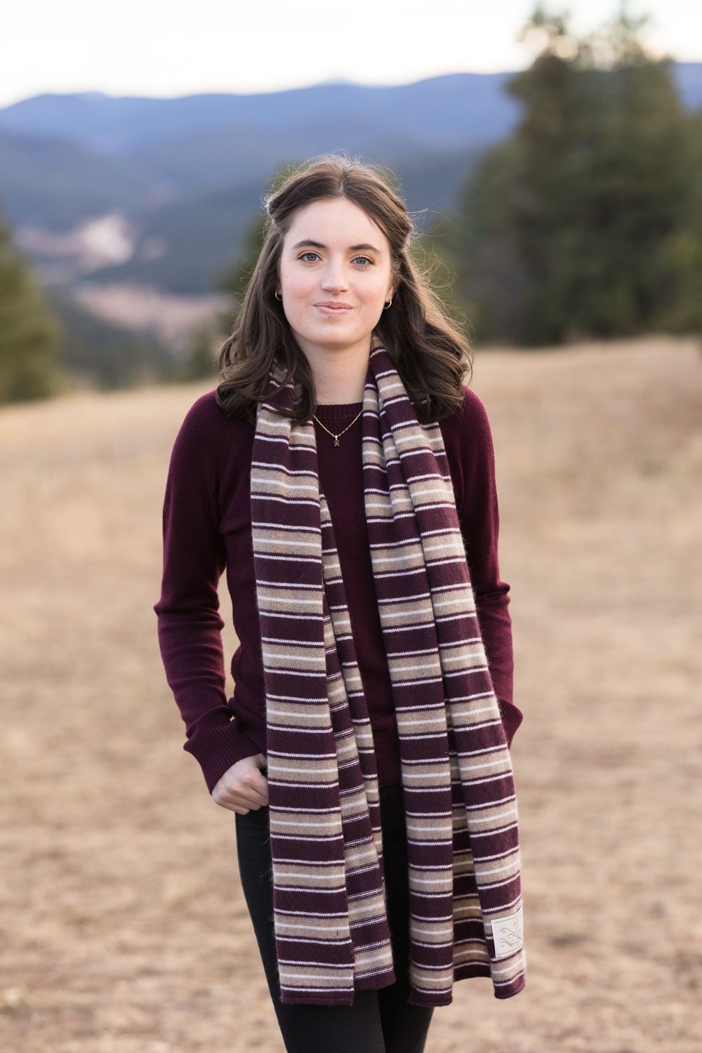 Person wearing a burgundy sweater and striped scarf poses outdoors with mountains in the background at sunset.