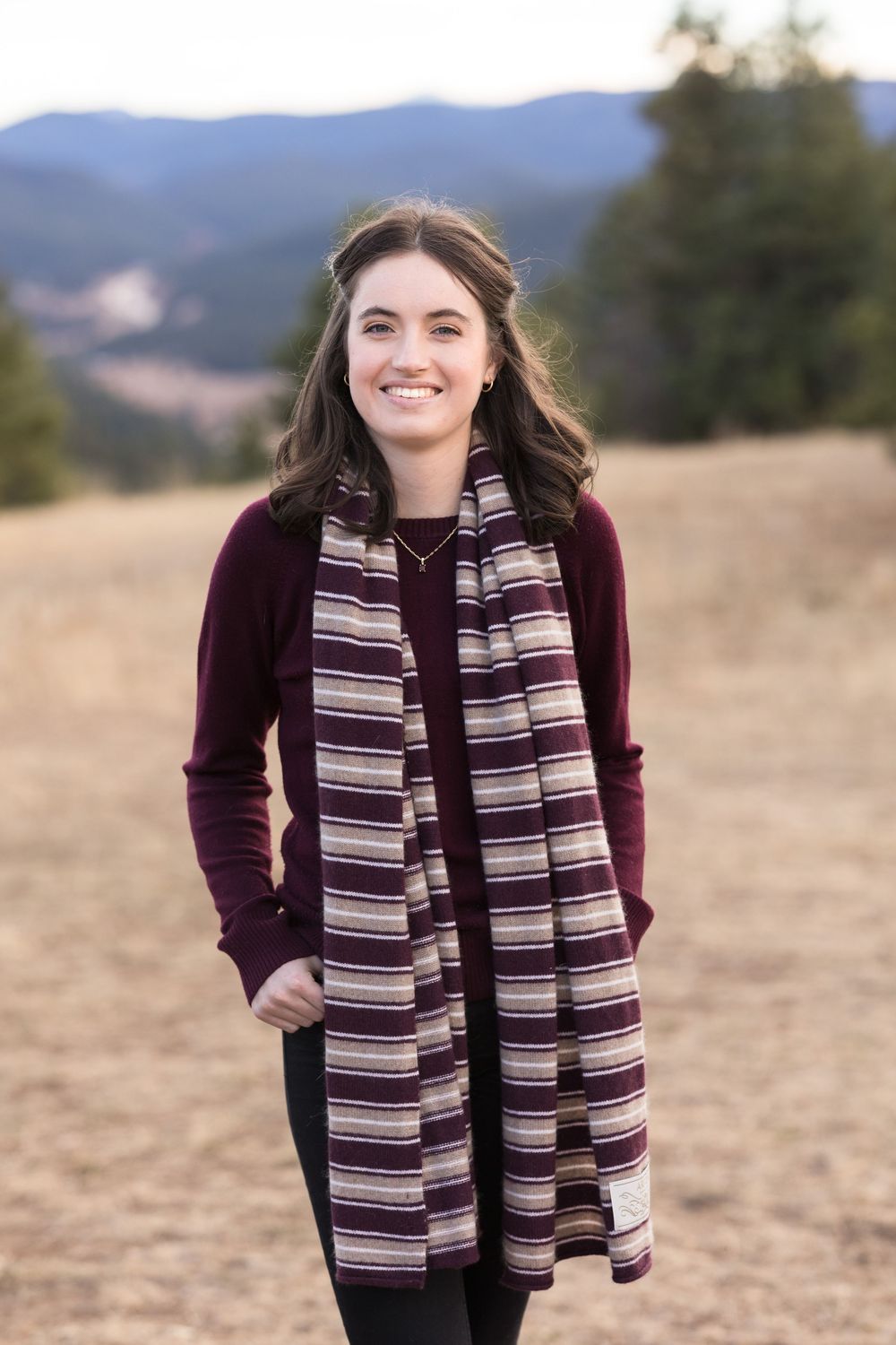 A person wearing a burgundy sweater and striped scarf poses outdoors in a field with mountains in the background.