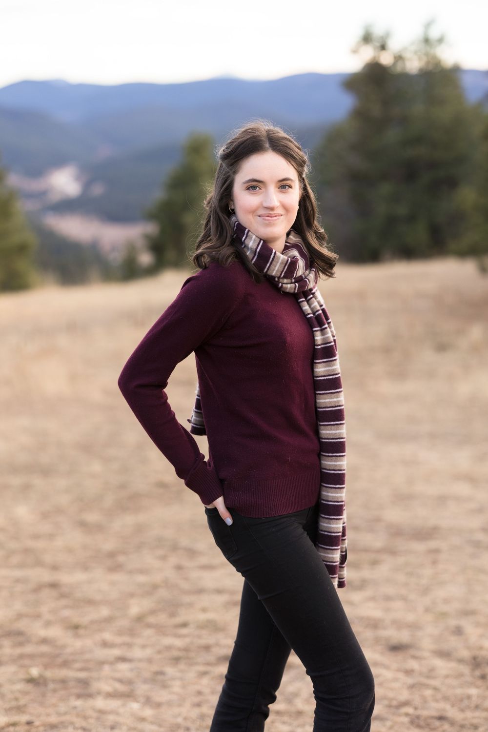 Person in burgundy sweater and striped scarf poses confidently in field with mountains behind.