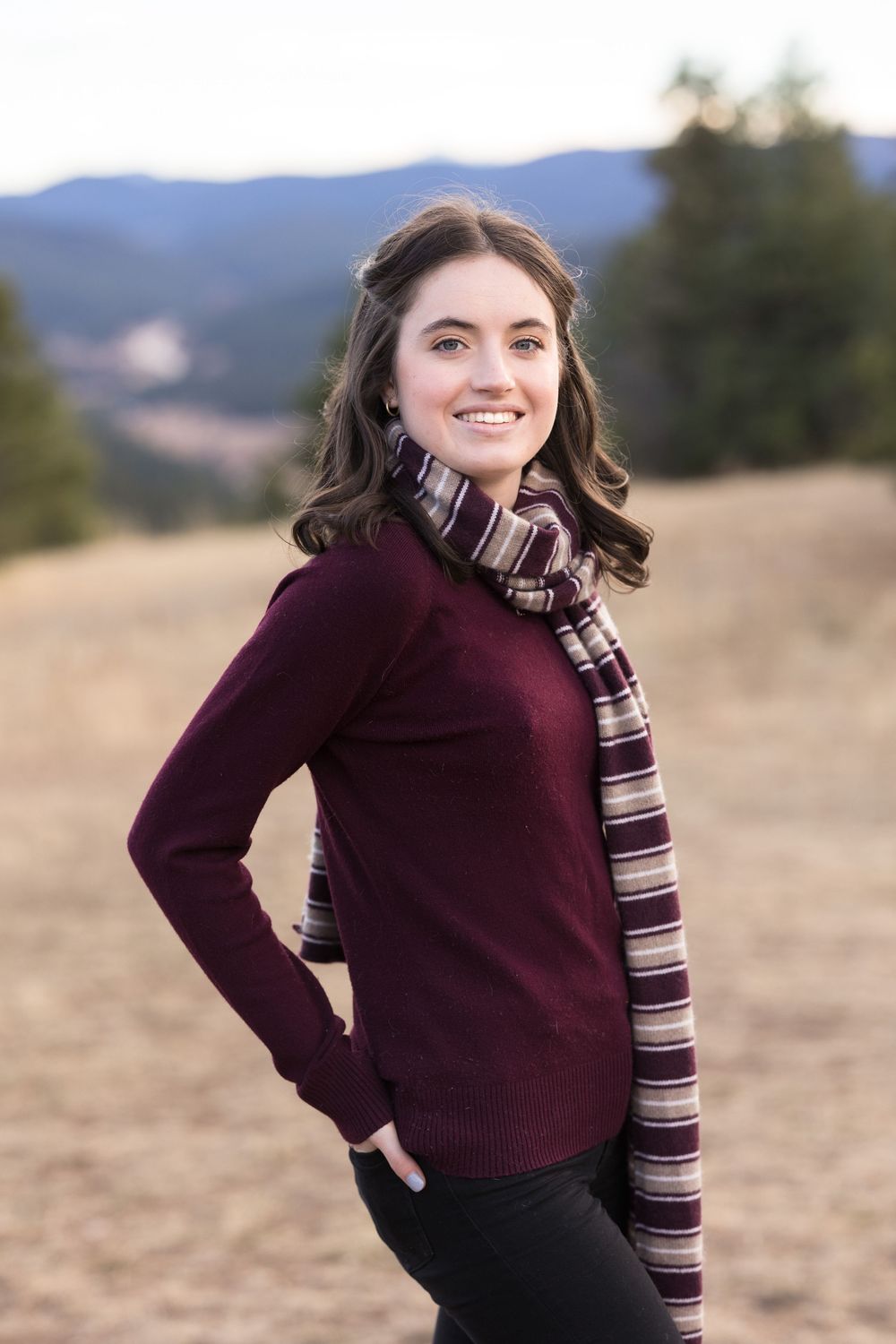 Portrait in a burgundy sweater and striped scarf with mountains in the background.
