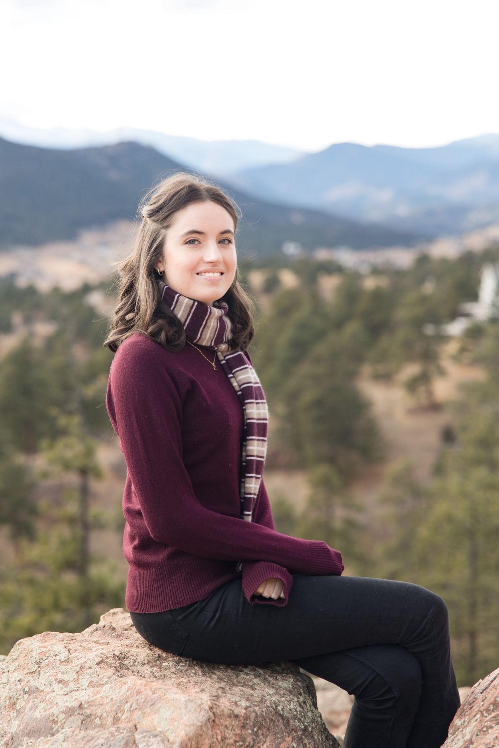 Person in burgundy sweater and plaid scarf sits on rocky overlook with mountain views in background.