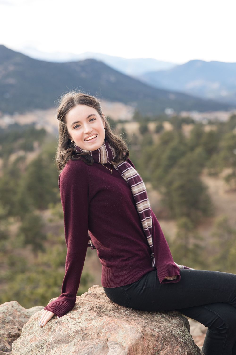 Person relaxes on rocky outcrop wearing maroon sweater against scenic mountain landscape.