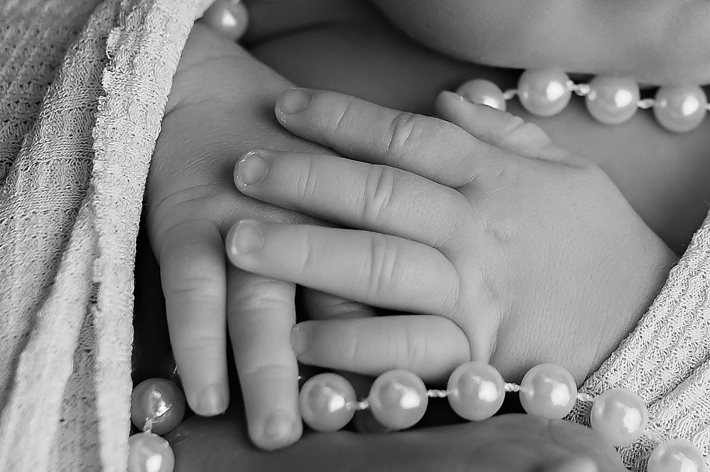 Black and white close-up of a baby's hand resting on chest with pearl necklace.