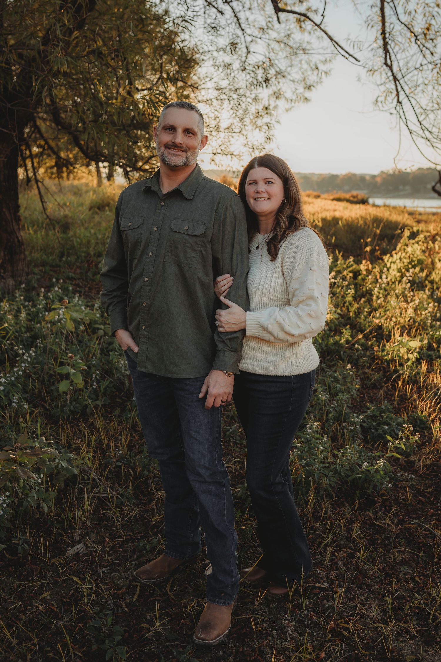 A couple stands together in a natural outdoor setting during golden hour with trees and water in the background.