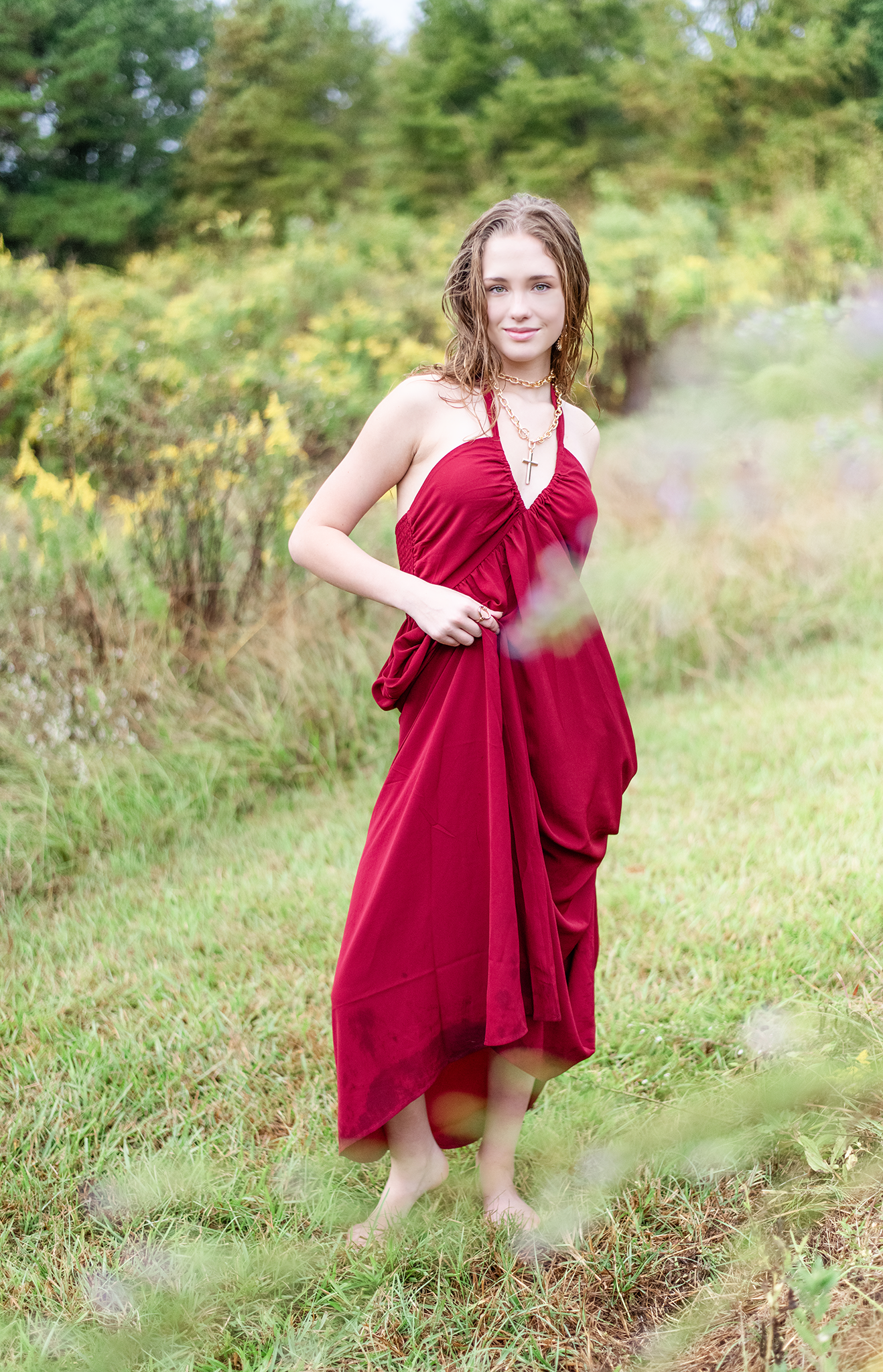 Young woman in elegant red halter dress standing barefoot in a lush green meadow.