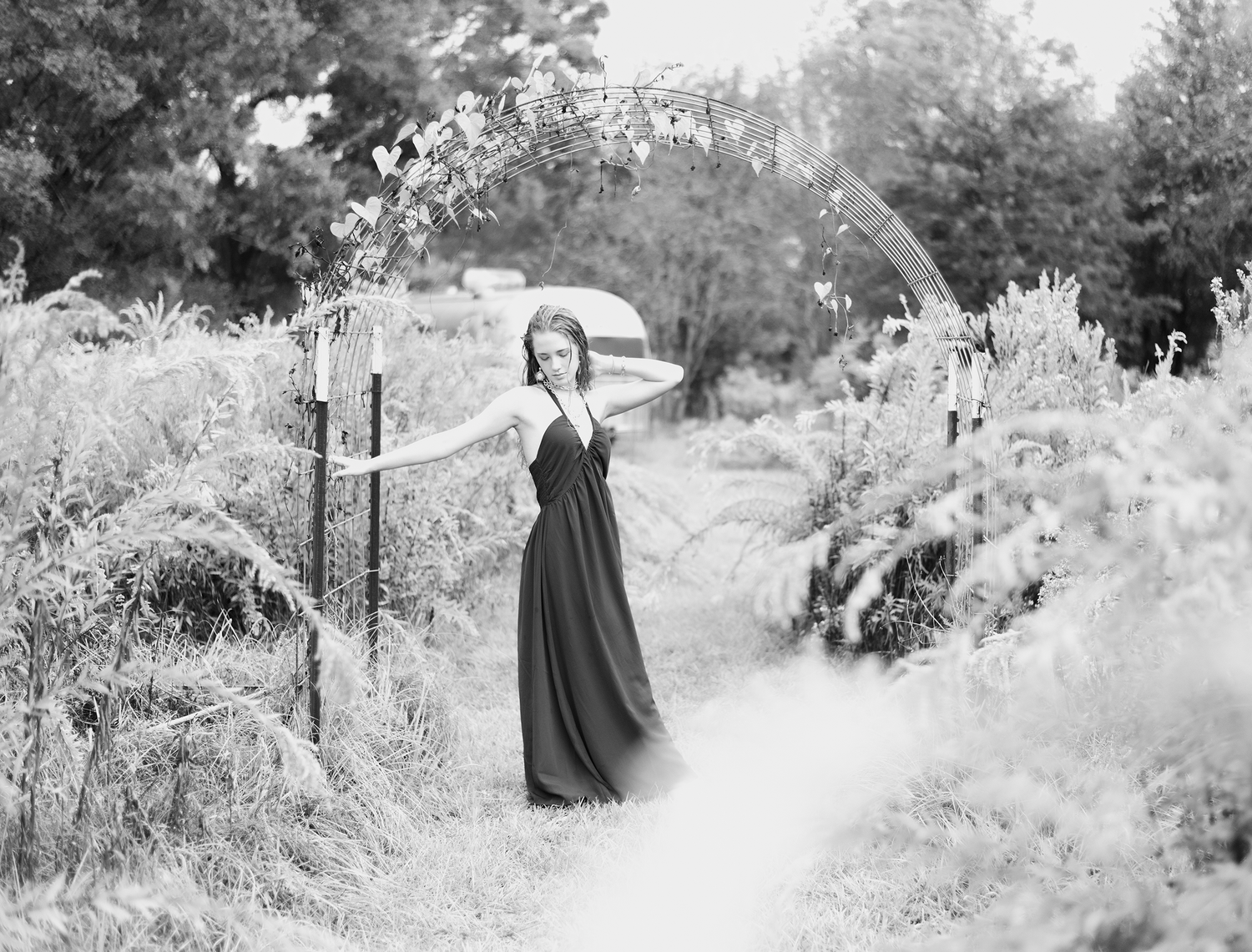 Black and white photo of a woman in a flowing gown posing under a floral arch outdoors.