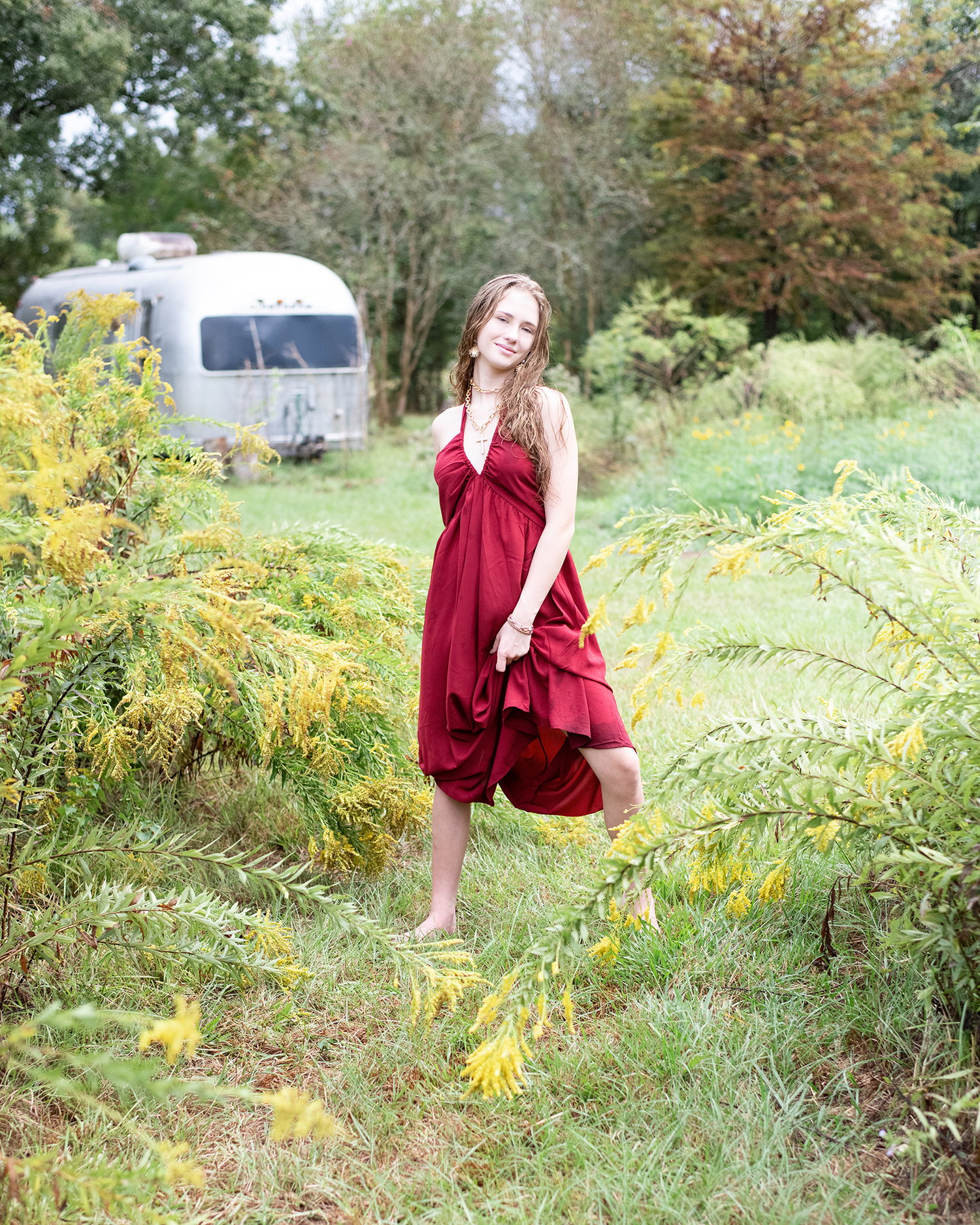 Young woman in red halter dress standing among yellow wildflowers with vintage Airstream trailer behind her.