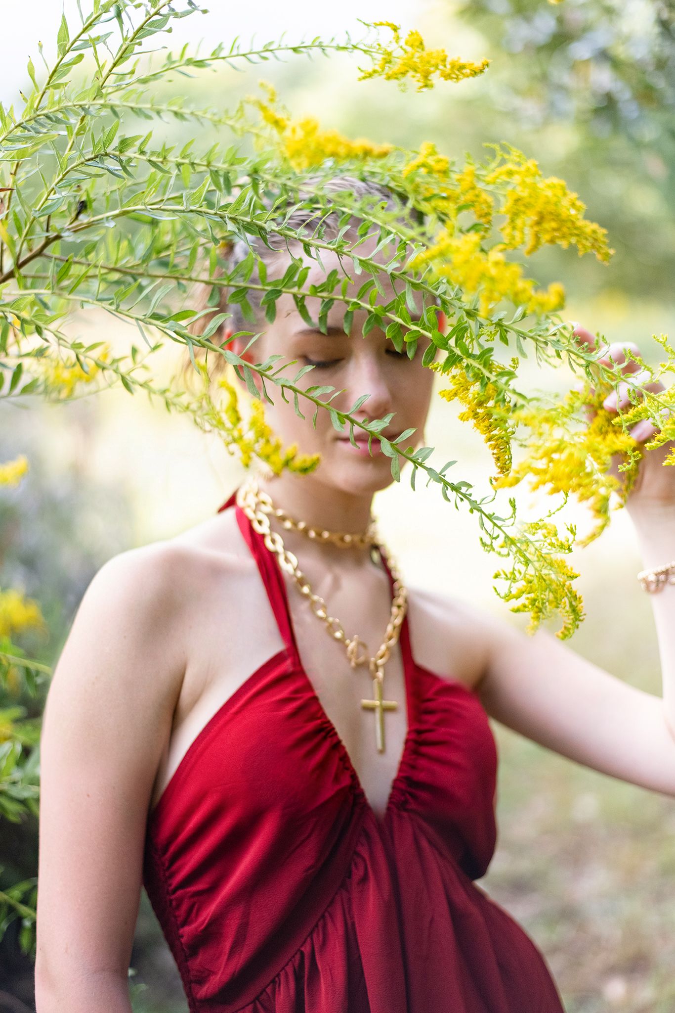 Young woman in red halter dress with eyes closed holding yellow wildflower branches over her face.