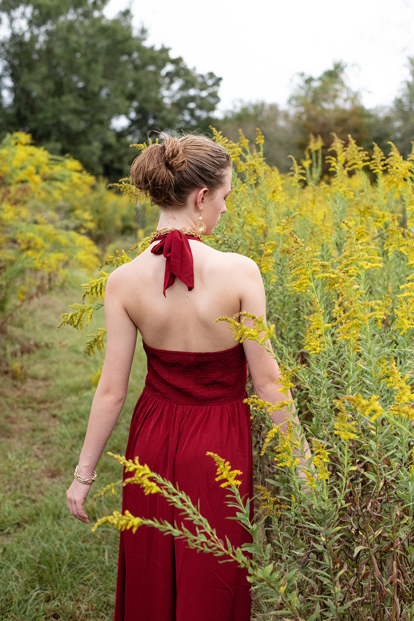 Woman in backless red halter dress walking through tall yellow goldenrod wildflowers in a field.