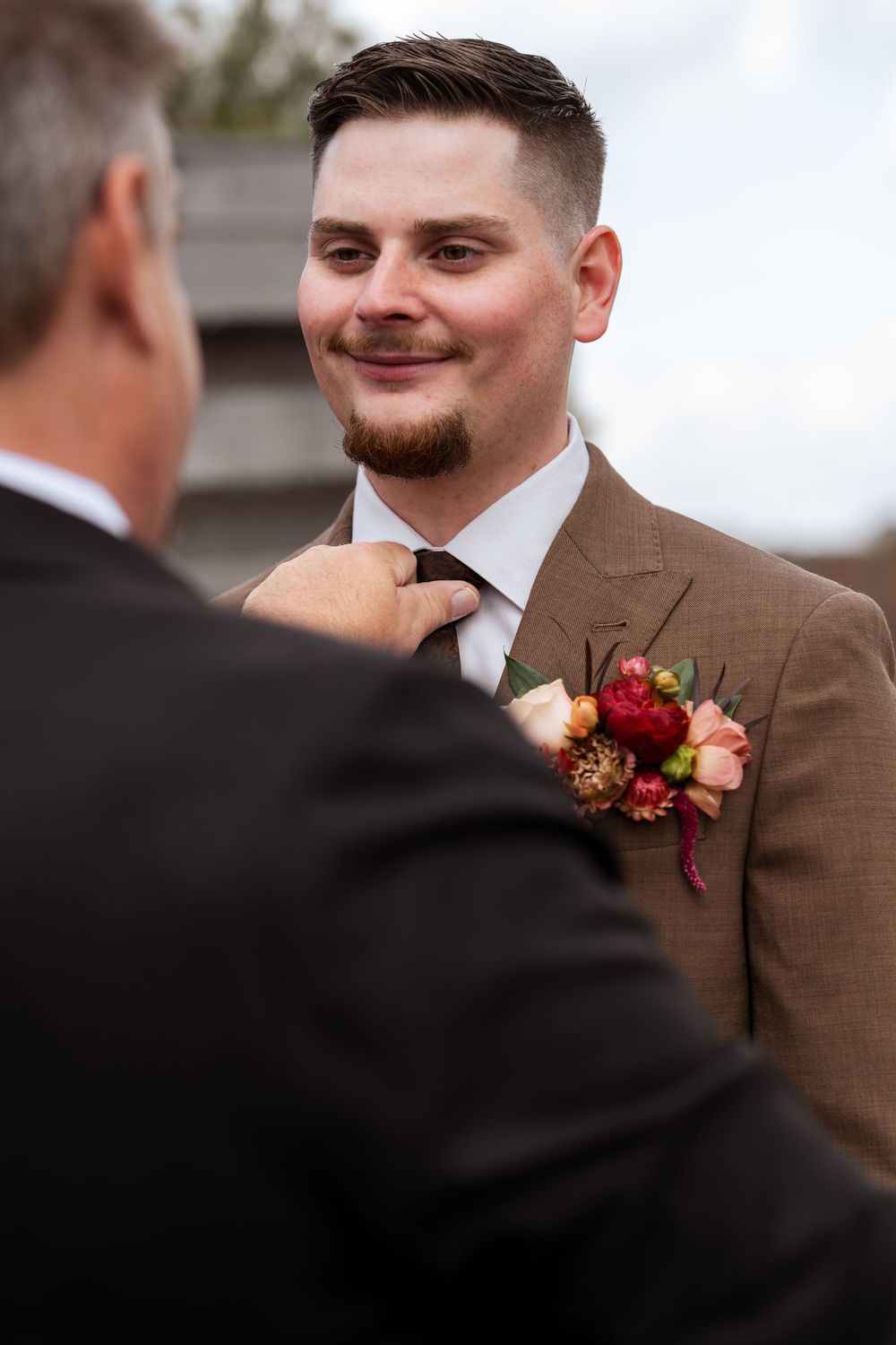 A groom in a brown suit with a red boutonniere has his tie adjusted during wedding preparations.