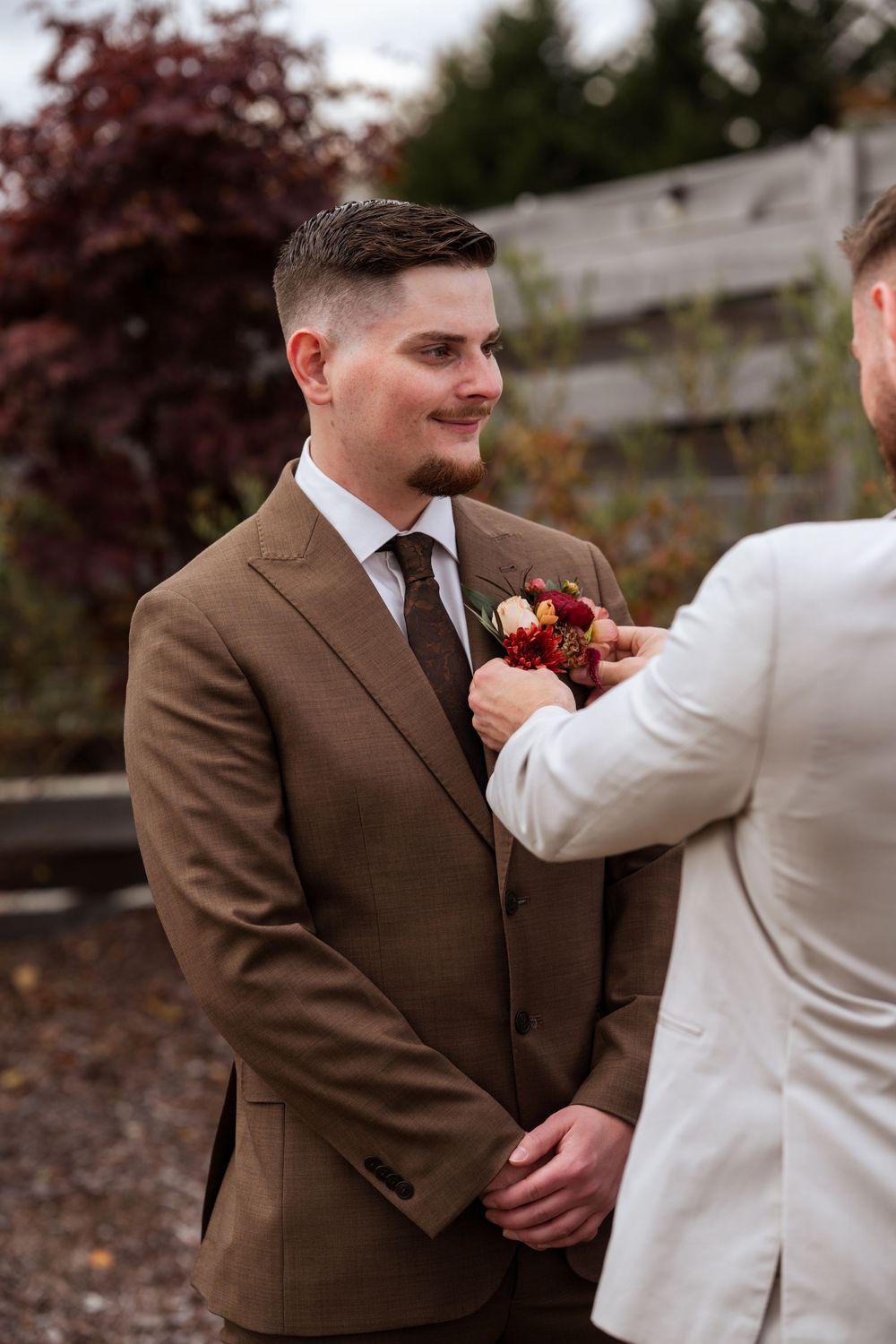 Wedding couple shares an intimate moment as the groom adjusts his boutonniere in a brown suit outdoors.