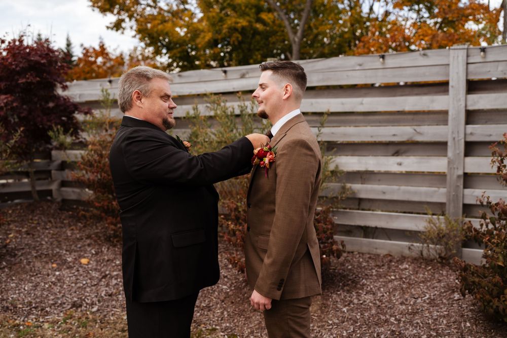 Wedding party members in brown suits interact outdoors by wooden fence during autumn.