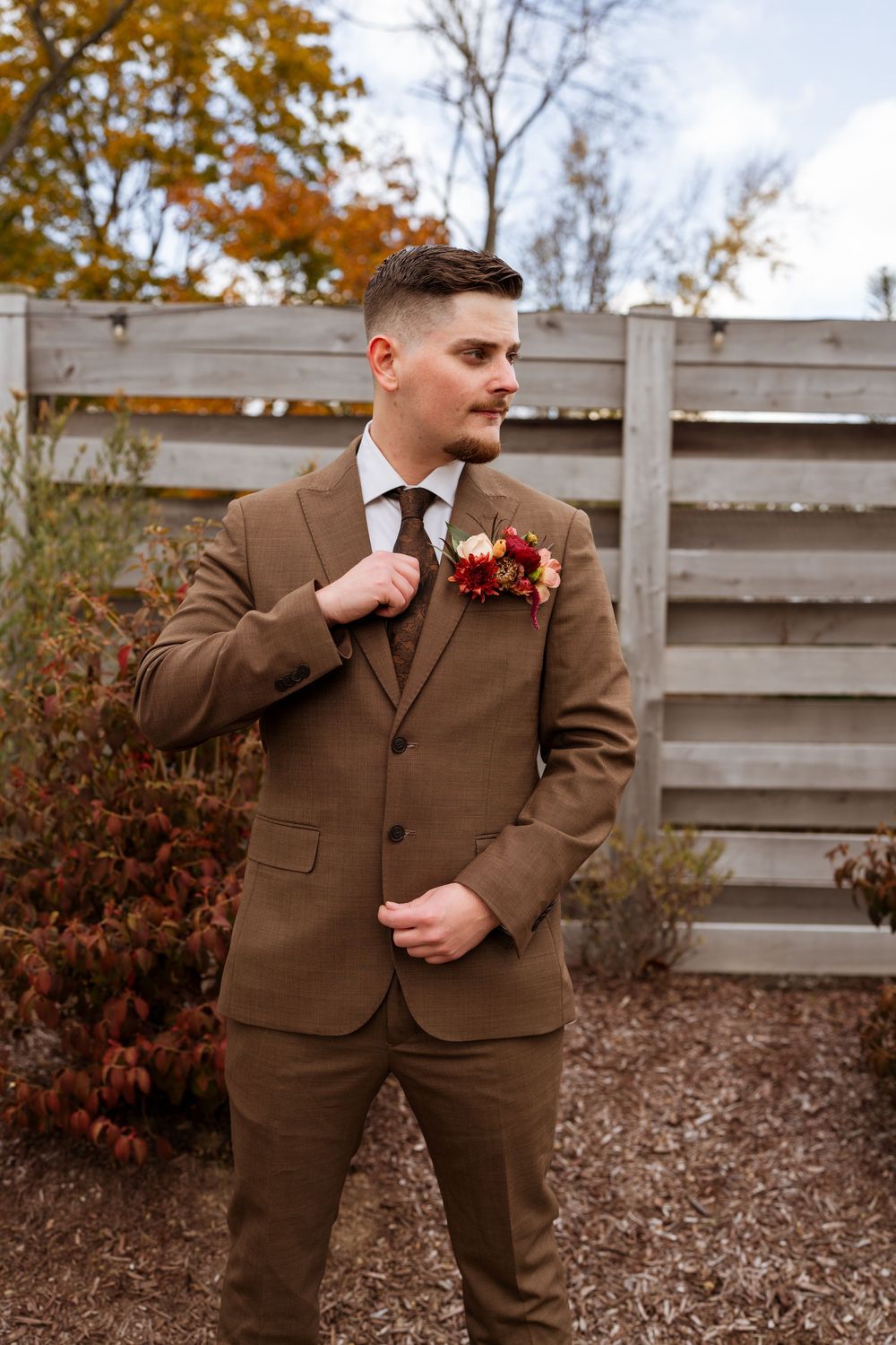 Groom in brown suit poses against wooden fence in autumn setting.