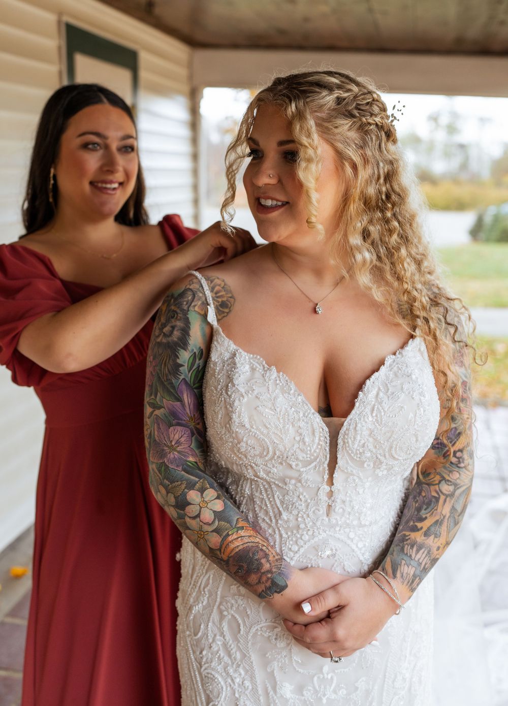 Bride in lace dress with tattoos poses with bridesmaids in burgundy dresses during wedding preparations.