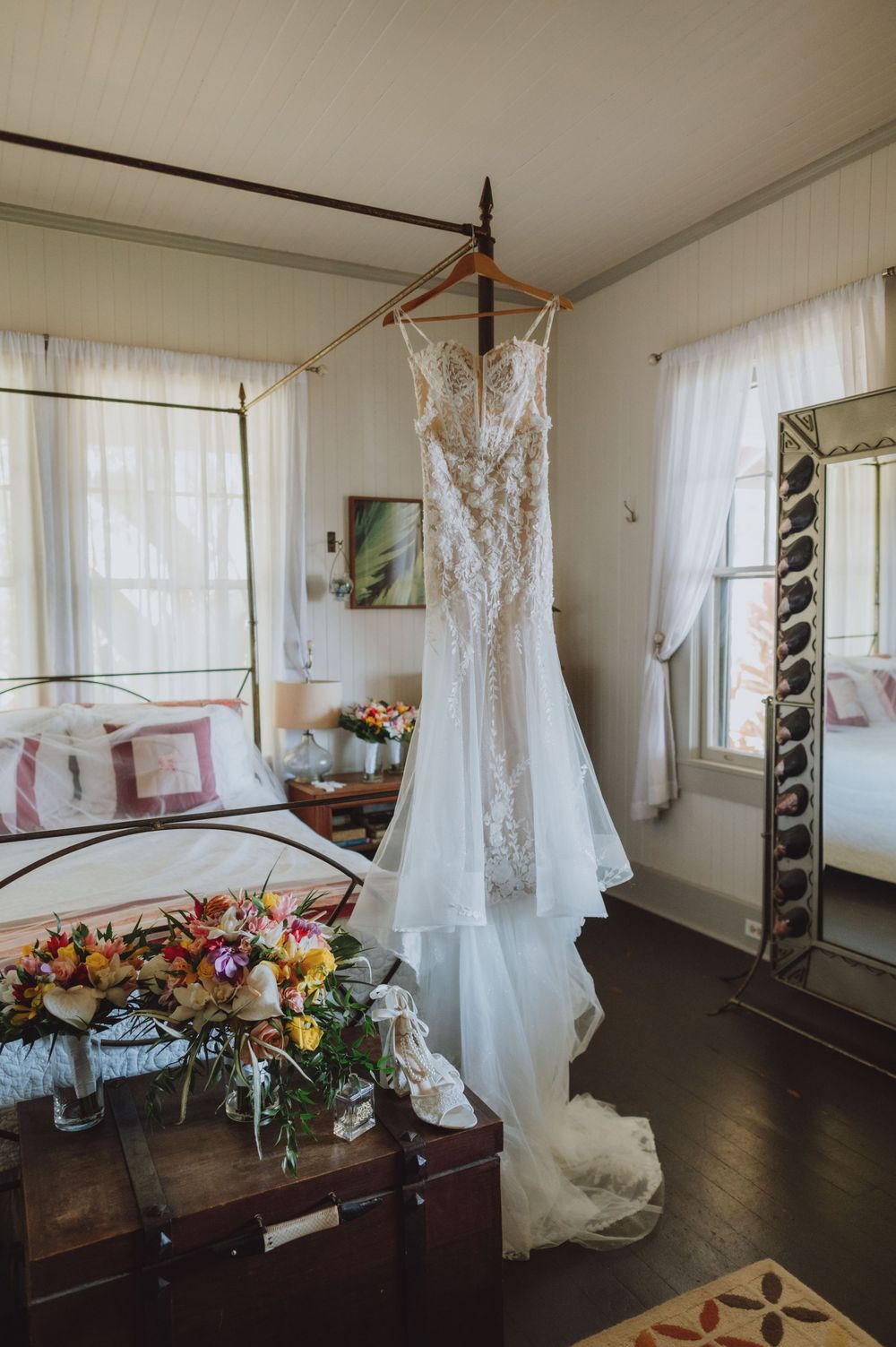 Wedding dress hanging by window in bridal suite with floral bouquet on wooden table.
