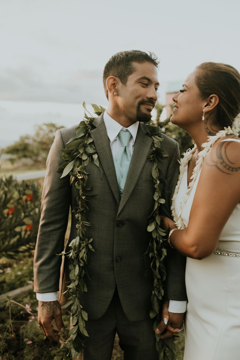 Couple wearing traditional wedding attire and green leaf leis share an intimate moment.